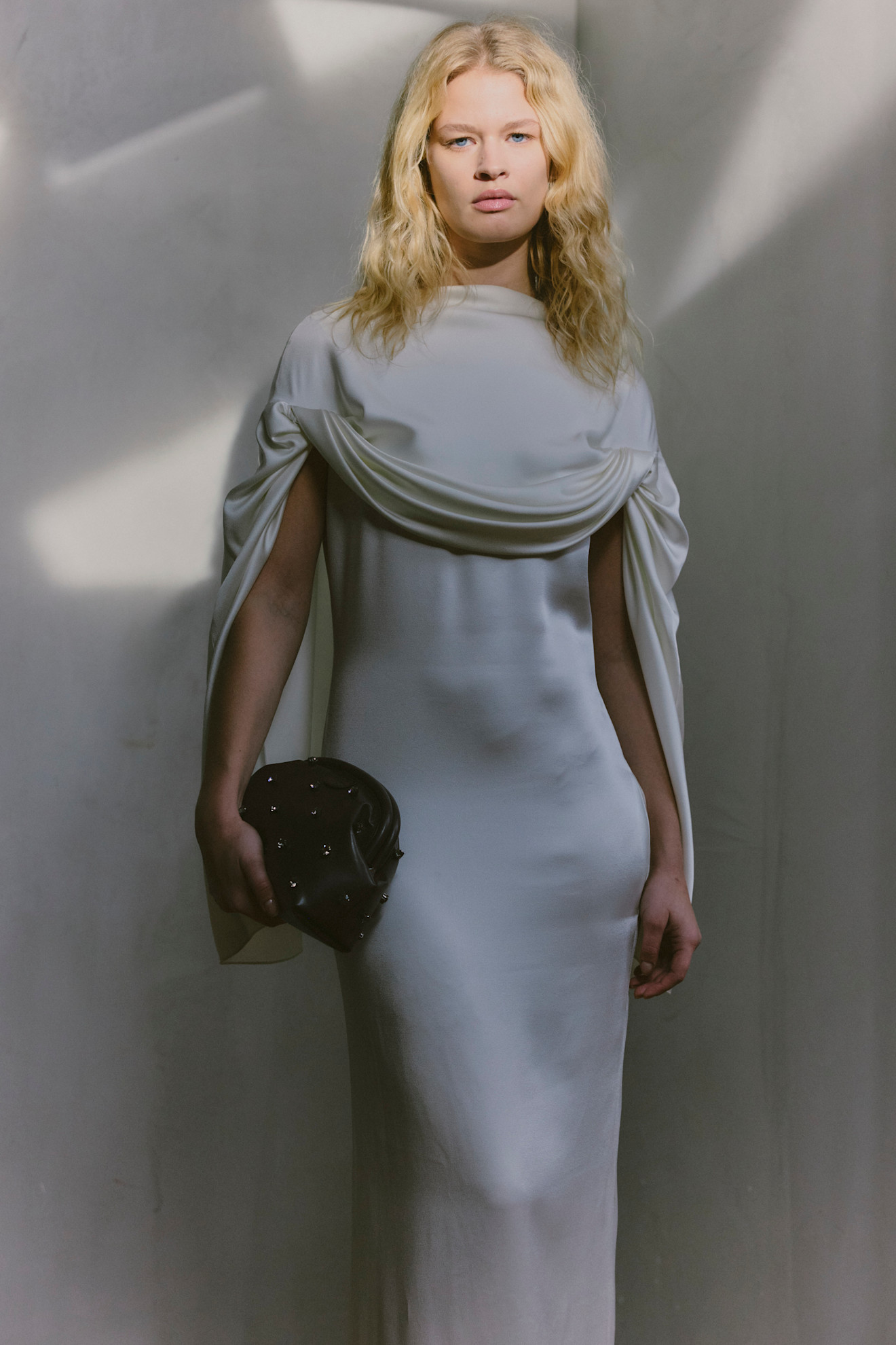 Обтягивающее белое платье с драпировкой на плечах, элегантный аксессуар в руках, мягкое освещение создает атмосферу утонченности