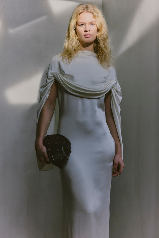 Обтягивающее белое платье с драпировкой на плечах, элегантный аксессуар в руках, мягкое освещение создает атмосферу утонченности