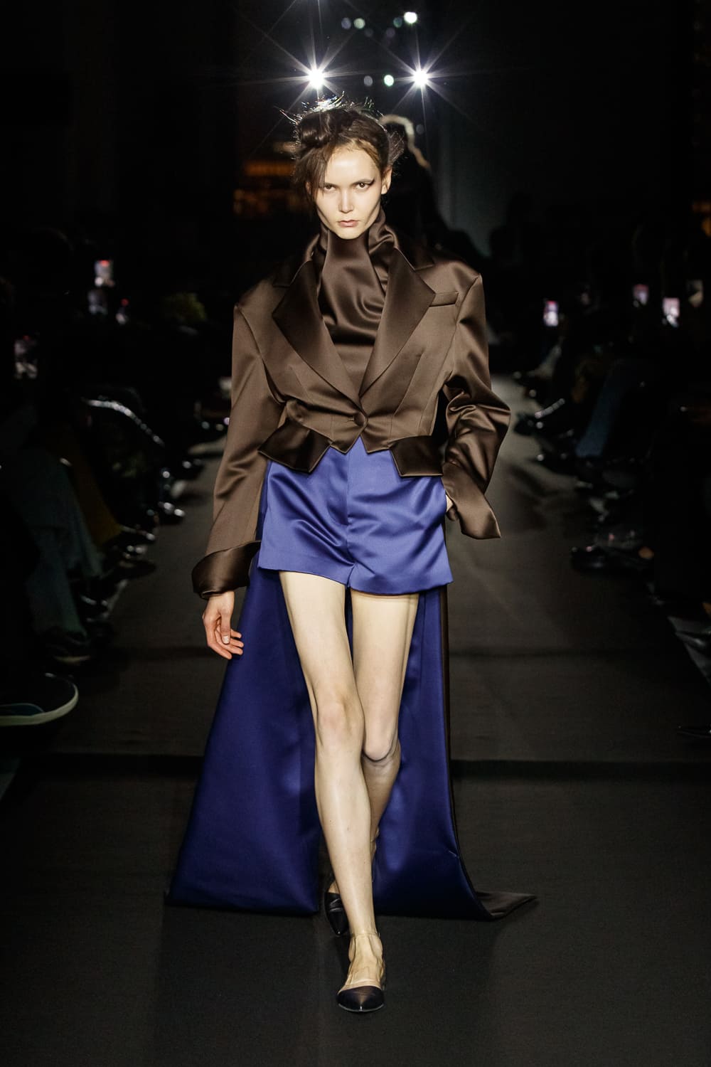 Модель в коричневом жакете и синем мини-юбке, создающая контраст с темным фоном подиума