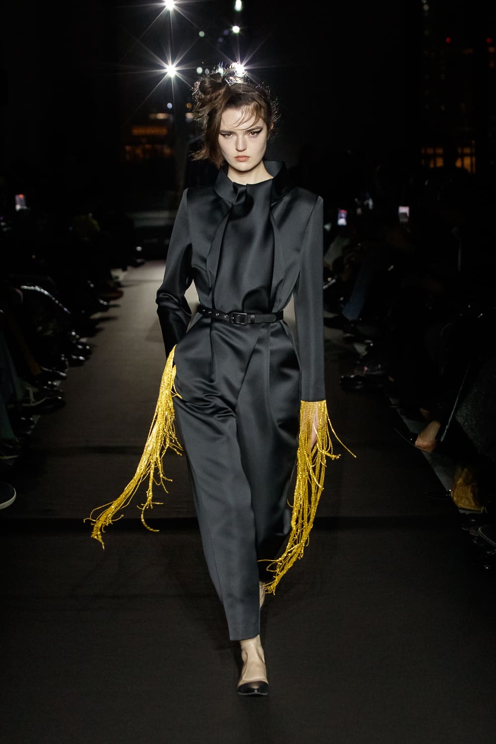 Элегантный черный комбинезон с золотыми бахромами, создающими эффект движения на фоне темного подиума