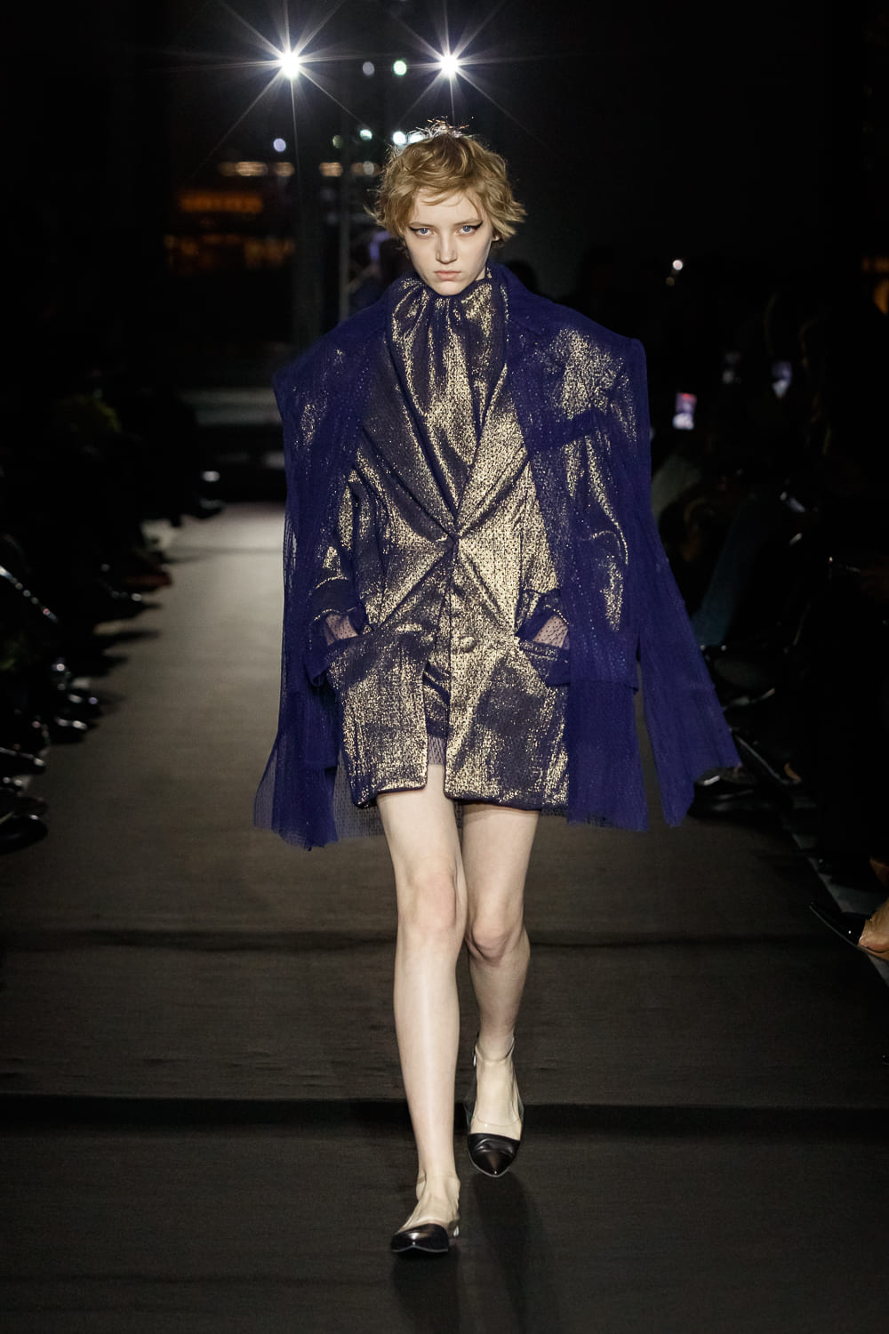 Модный наряд с золотистым жакетом и прозрачным капюшоном, создающий контраст с темным фоном подиума