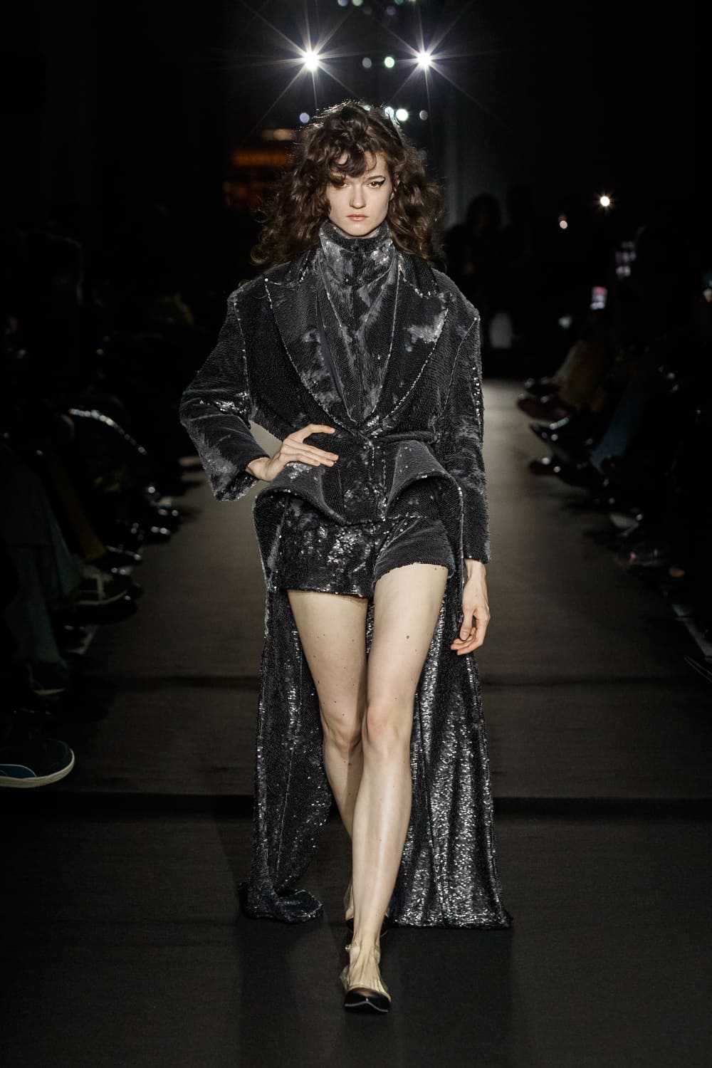 Блестящий наряд с короткими шортами и длинным пиджаком, создающий эффект вечернего сияния на подиуме
