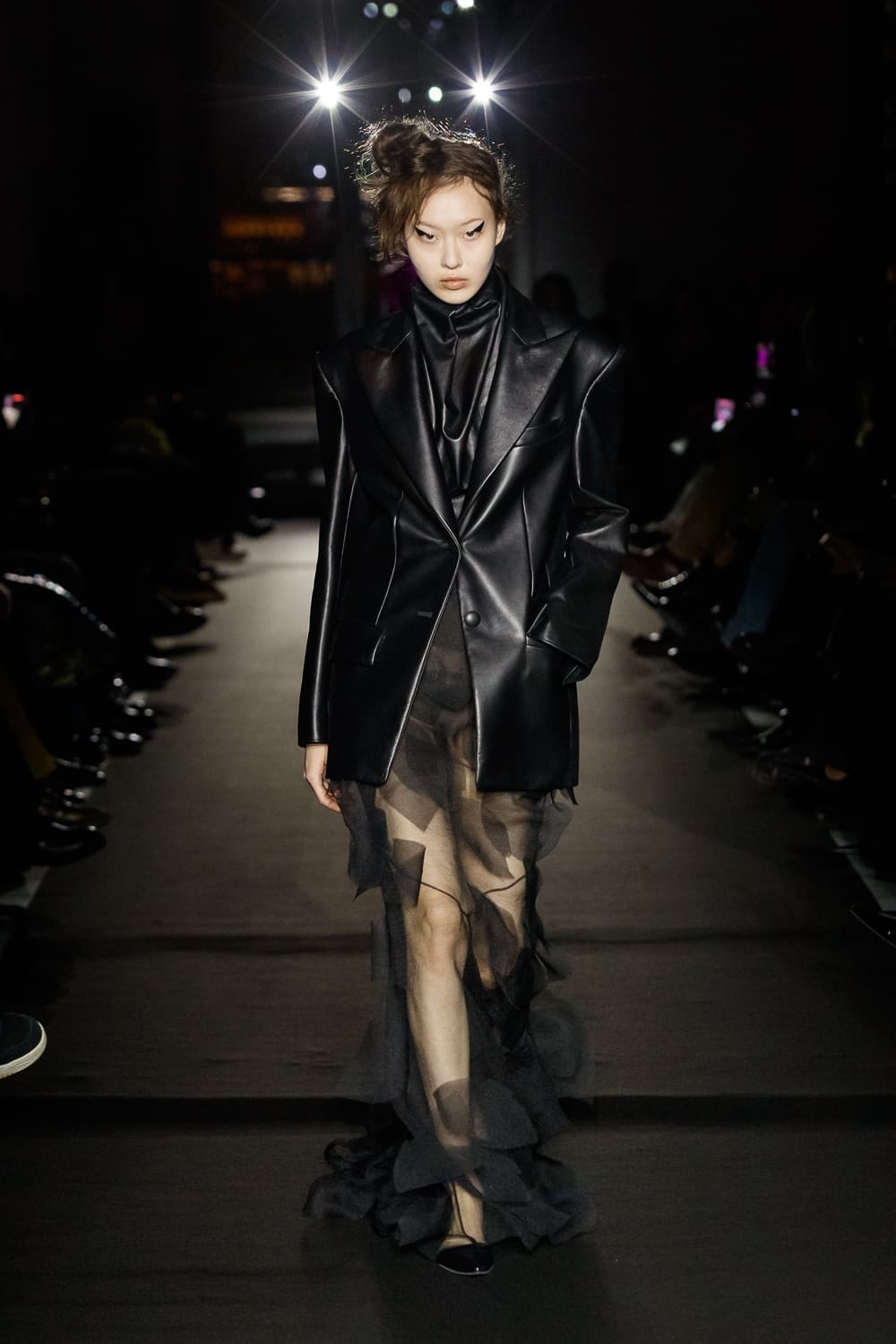 Образ в черном кожаном пиджаке с прозрачной юбкой, создающий эффект легкости и загадочности на подиуме