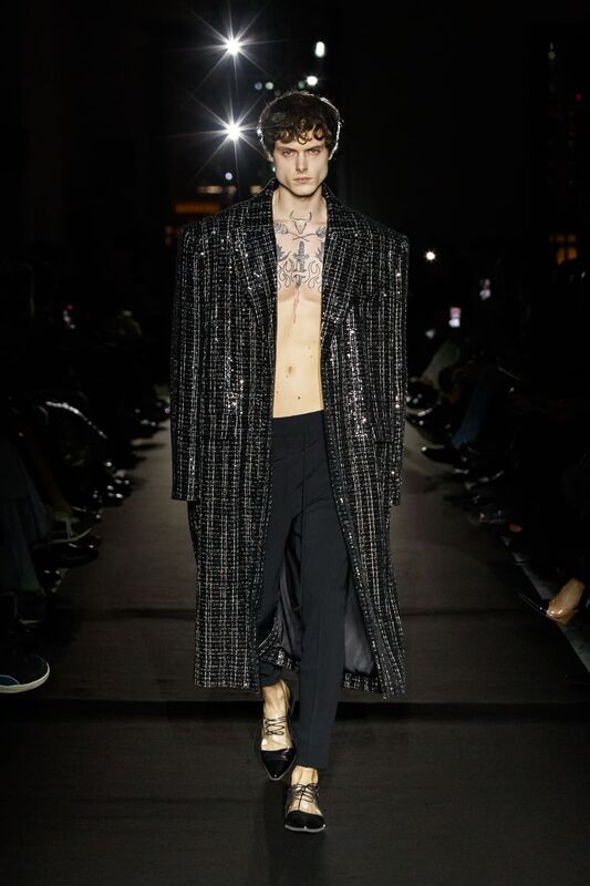 Мужской образ в длинном блестящем пальто с открытой грудью и черными брюками, сочетание стиля и дерзости на подиуме.