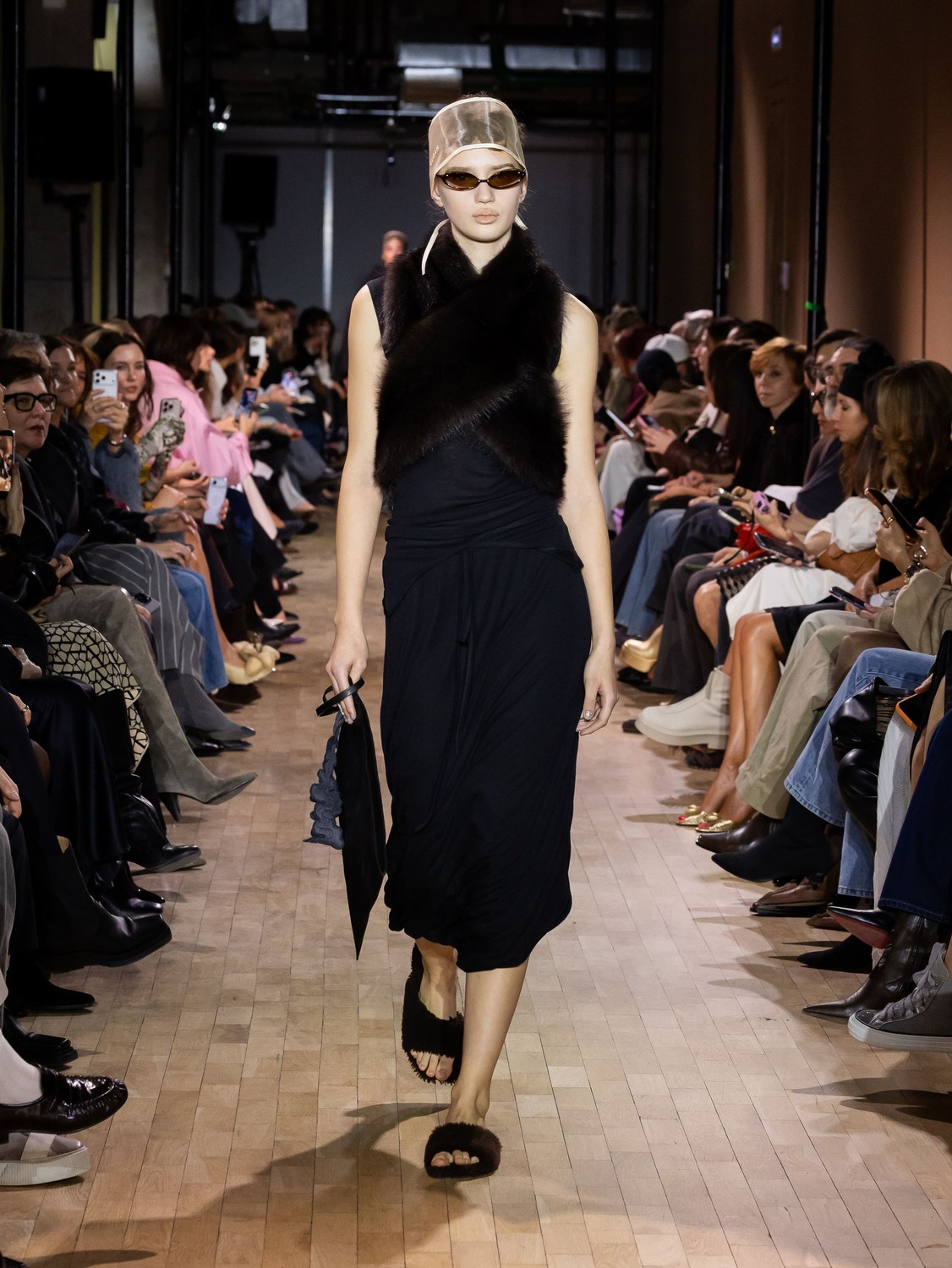 Черное платье с асимметричным кроем и меховым шарфом, дополненное стильными очками и сандалиями, создает загадочный образ