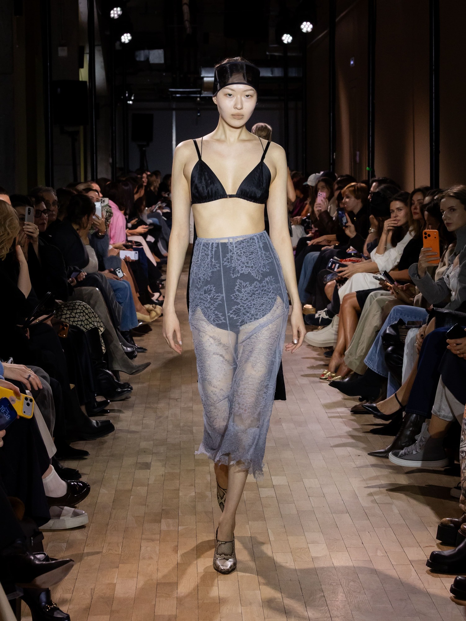 Модель в черном бюстгальтере и полупрозрачной юбке с цветочным узором, создающей игривый контраст с минималистичным стилем показа