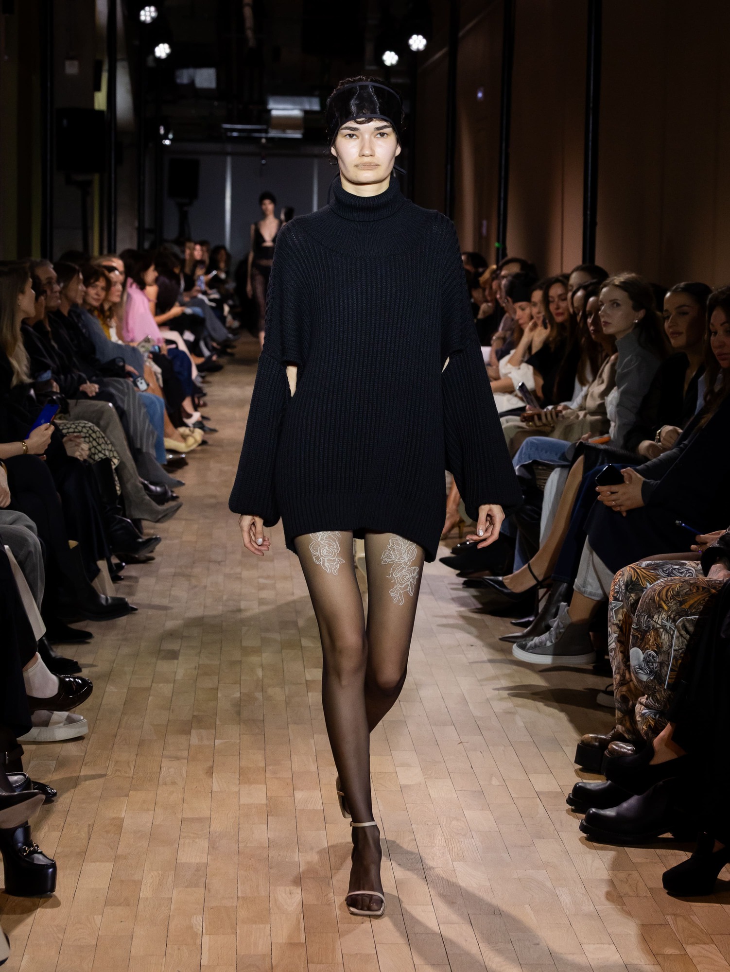 Объемный черный свитер с широкими рукавами, дополненный прозрачными колготками с цветочным узором, создает эффектный контраст на подиуме
