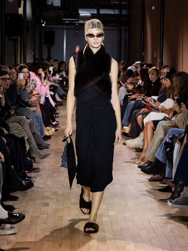 Черное платье с асимметричным кроем и меховым шарфом, дополненное стильными очками и сандалиями, создает загадочный образ