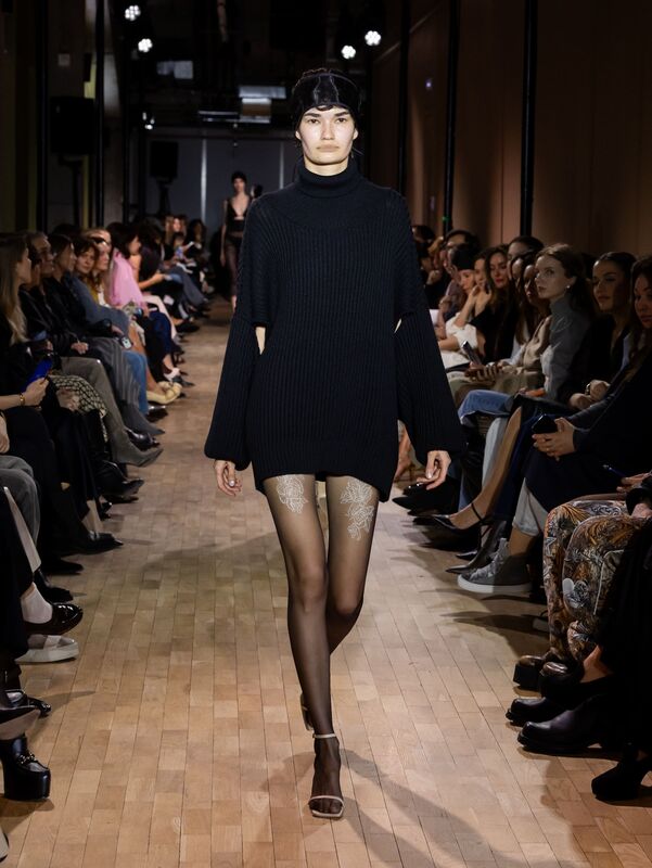 Объемный черный свитер с широкими рукавами, дополненный прозрачными колготками с цветочным узором, создает эффектный контраст на подиуме