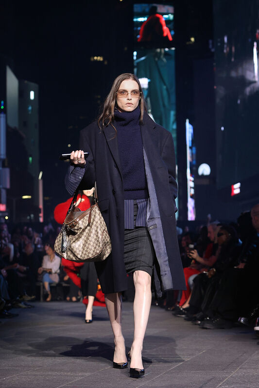 Модель в длинном темном пальто и свитере, сочетает строгую юбку и сумку Gucci, создавая атмосферу городской элегантности